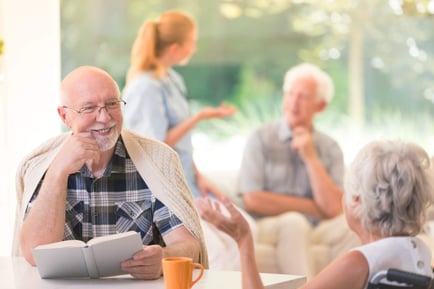 An older adult smiles warmly while holding a book and chatting in a bright, sunlit common area as others converse nearby, capturing the engaged, connected lifestyle of senior living Herndon Virginia at Arbor Terrace Herndon, a vibrant independent living Herndon VA community