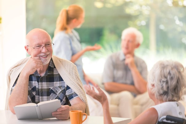 An older adult smiles warmly while holding a book and chatting in a bright, sunlit common area as others converse nearby, capturing the engaged, connected lifestyle of senior living Herndon Virginia at Arbor Terrace Herndon, a vibrant independent living Herndon VA community