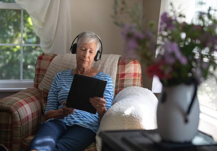 An older adult relaxes in a cozy armchair wearing headphones and browsing a tablet beside a sunny window and fresh flowers, reflecting the comfortable, independent senior lifestyle Herndon VA residents enjoy at Arbor Terrace Herndon.
