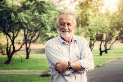 A cheerful older gentleman with a warm smile stands outdoors among lush green trees, representing the active senior lifestyle available through senior living options in Union County, New Jersey.