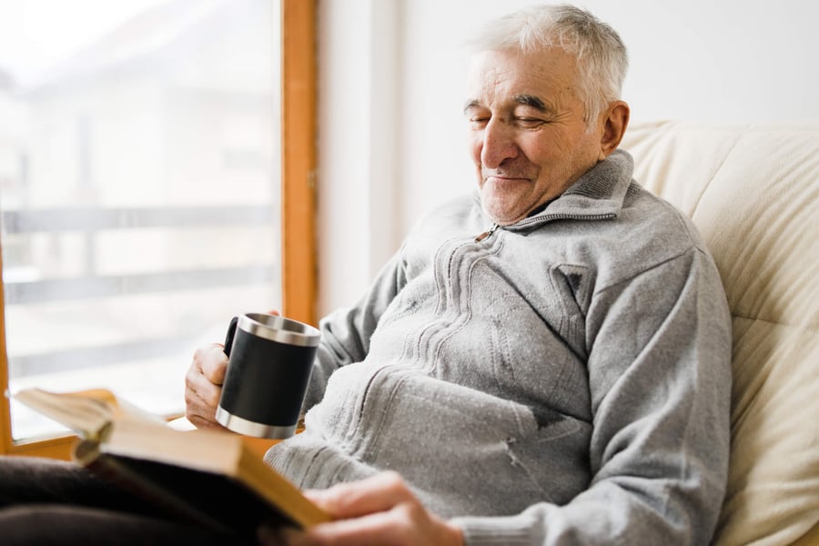 An older adult relaxes contentedly in a cozy chair with a warm mug and a book by a bright window, capturing the comfortable, peaceful lifestyle of senior living in Morris Plains, NJ.