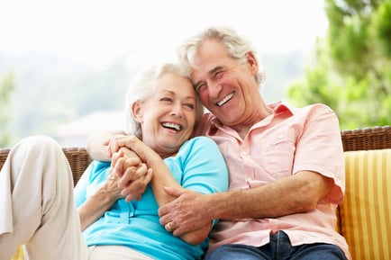 Two older adults snuggle and laugh together on an outdoor seat surrounded by greenery, reflecting the warm, loving connections and wellness-focused senior living experience at Arbor Terrace Morris Plains, NJ.