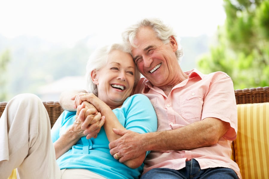 Two older adults snuggle and laugh together on an outdoor seat surrounded by greenery, reflecting the warm, loving connections and wellness-focused senior living experience at Arbor Terrace Morris Plains, NJ.