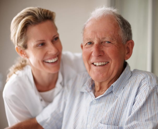 A younger woman and an older man share warm smiles side by side in a softly lit setting, reflecting the caring, person-centered approach to senior living in Athens, GA.