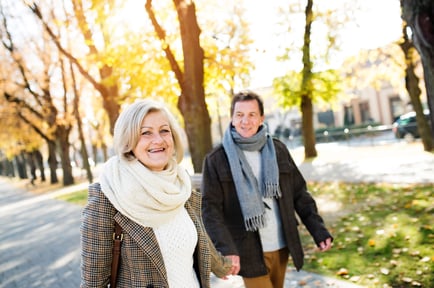Two seniors smile and walk together along a tree-lined path during autumn, enjoying the active senior lifestyle that makes Mountainside, NJ a wonderful place for senior living.