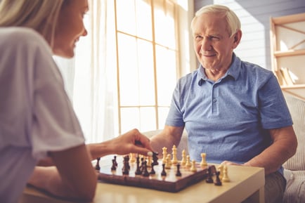 An older man smiles warmly while playing chess with another person near a sun-filled window, enjoying a mentally engaging activity at a senior living community in Athens, GA.