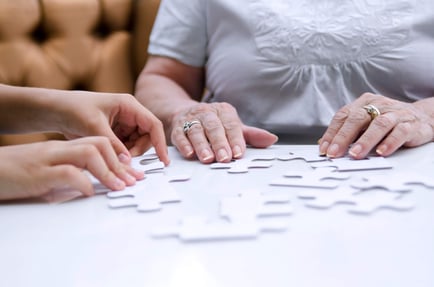 Two sets of hands gently work together on a white puzzle at a memory care community in Fort Myers, FL, a calming, engaging activity that supports connection and cognitive wellness for residents.