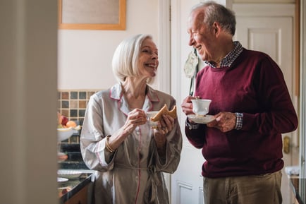 Two older adults share a cheerful morning moment in a home kitchen, enjoying tea and a light snack together — reflecting the warm, maintenance-free senior living Herndon lifestyle offered at Arbor Terrace Herndon, a top independent living Herndon VA community.