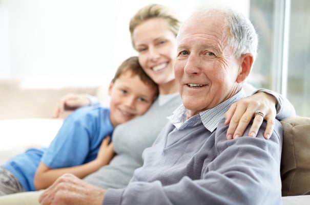 An older adult smiles warmly while being embraced by a younger adult and a child on a sofa — a tender family moment supporting early-stage dementia care near Bee Cave TX.