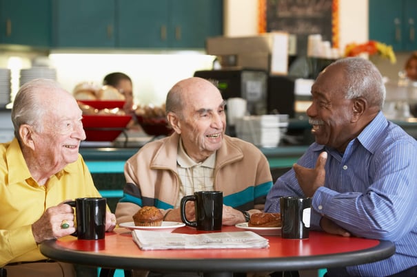 Three older adults laugh together over coffee and muffins at a casual dining counter — the warm social connections found at assisted living Lakeway Texas.