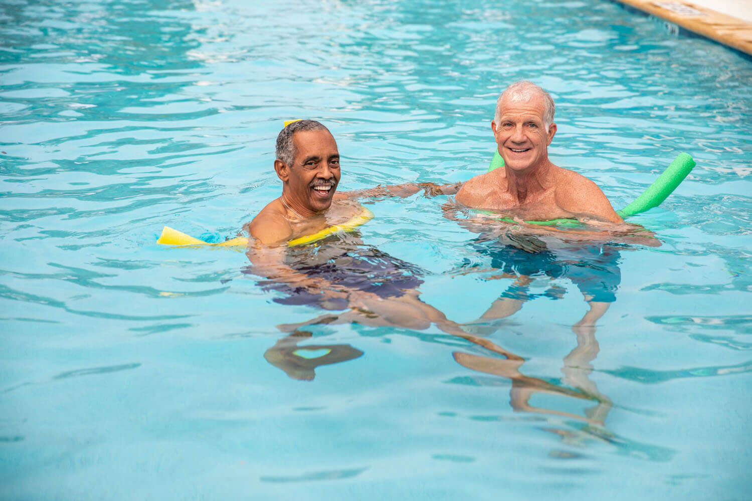 Two smiling senior men float in a sparkling blue pool with colorful foam noodles, enjoying a sunny swim together, a refreshing moment that captures active senior living Atlanta, GA and top-rated senior living in Atlanta, GA.