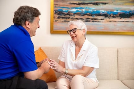 Smiling resident in glasses and a white blouse holds hands with a team member in a blue polo as they talk together on a cream sofa beneath a colorful abstract painting.