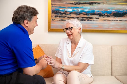 Smiling resident in glasses and a white blouse holds hands with a team member in a blue polo as they talk together on a cream sofa beneath a colorful abstract painting.