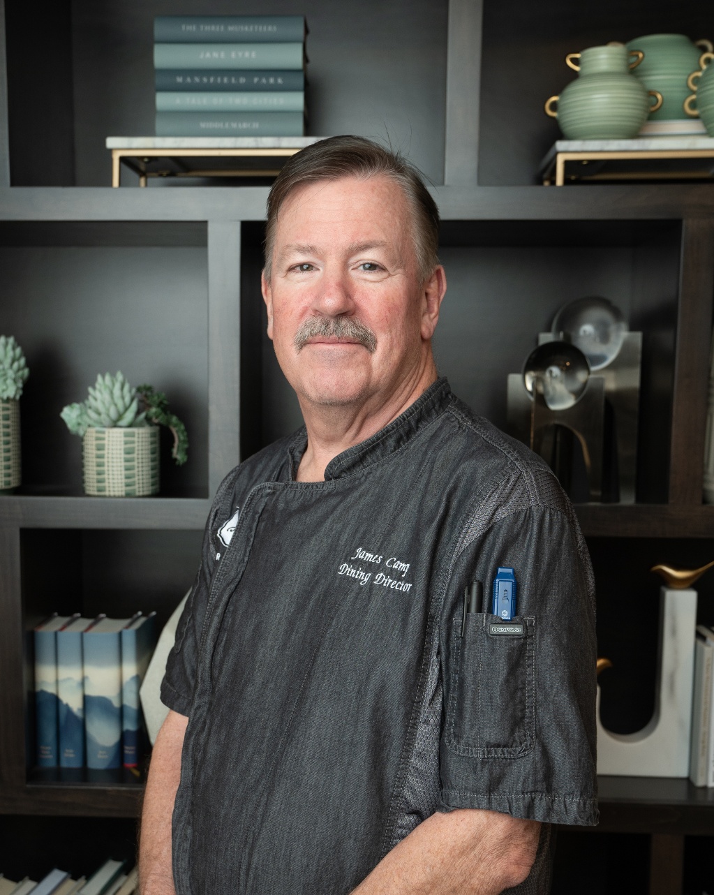 James Camp, Dining Director at Arbor Terrace Highland Park, smiles in a dark gray chef coat in front of shelves with a potted succulent and decor, leading a dining experience that reflects resort-style senior living Lake County IL and an active senior lifestyle Highland Park IL.