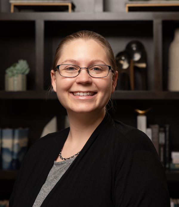 Kate Kloman, Memory Care Director at Arbor Terrace Highland Park, smiles in glasses and a black cardigan in front of bookshelves, part of a caring team at one of the top senior living communities Lake County IL offering personalized senior care Highland Park IL.