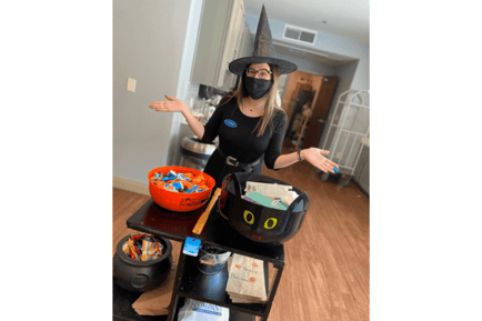 A staff member in a witch costume and mask stands beside a table of Halloween candy bowls — fall community festivities at Arbor Terrace Lakeway senior living Texas.
