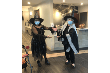 Two people in witch costumes and masks strike a playful pose in a warmly lit lobby decorated for Halloween — festive community events at assisted living Lakeway TX.