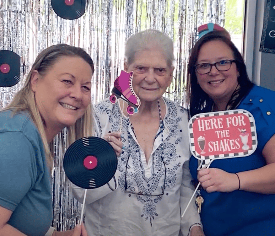 Three people smile together at a sock hop-themed photo booth with vinyl record props at Arbor Terrace Lakeway — joyful senior living Lakeway Texas with engaging community events.