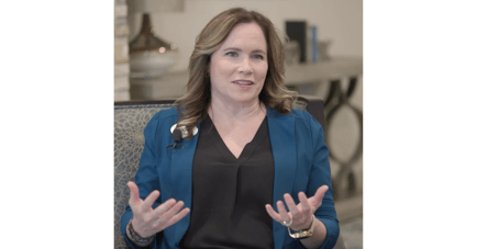 A woman in a blue blazer speaks expressively in a warmly furnished room — sharing expert guidance on dementia care Lakeway TX and how to help loved ones cope and thrive.