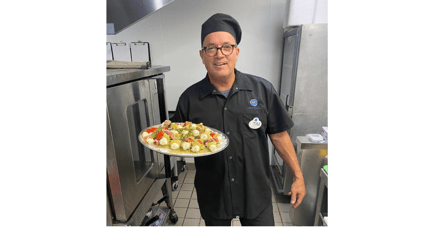A smiling staff member in a chef's uniform holds a platter of freshly prepared appetizers in the kitchen — fresh, chef-crafted dining at senior living Lakeway Texas.