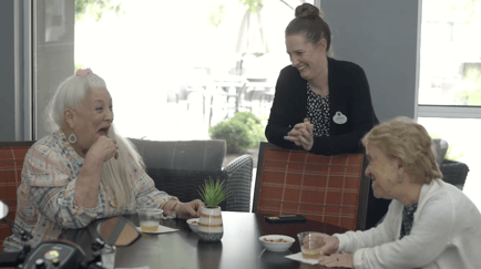 A staff member shares a warm, laughing moment with two residents at a dining table at Arbor Terrace Lakeway — senior care Lakeway TX defined by joy and meaningful relationships.