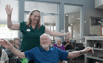A staff member guides a smiling older adult through a stretching exercise while other seniors look on — active senior lifestyle and wellness-focused assisted living Lakeway TX.