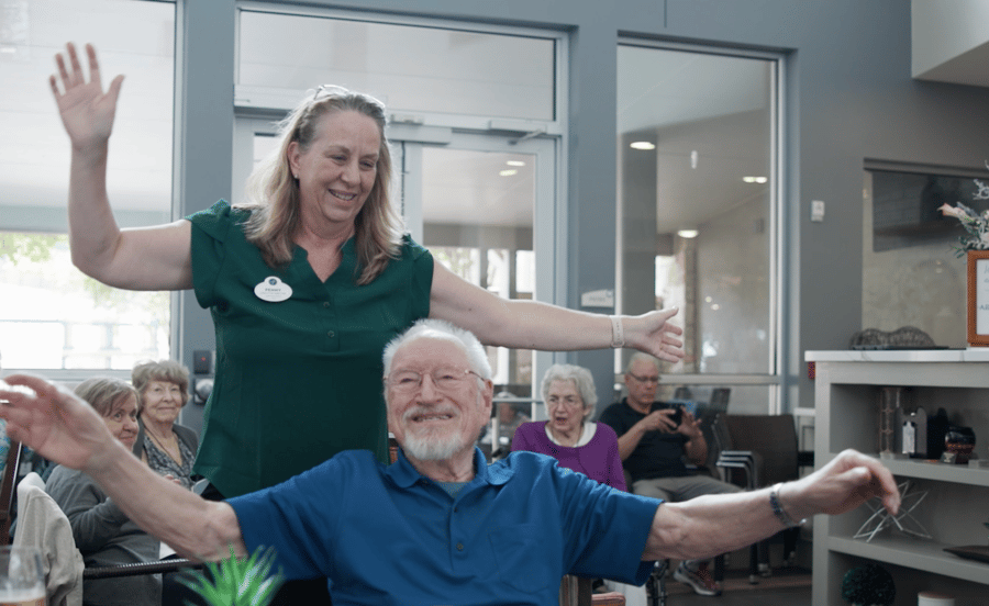A staff member guides a smiling older adult through a stretching exercise while other seniors look on — active senior lifestyle and wellness-focused assisted living Lakeway TX.