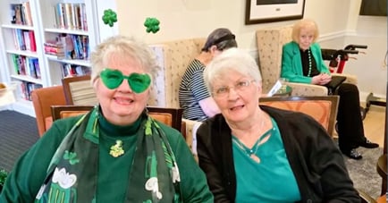 Two elderly women smiling together at a St. Patrick's Day celebration, both dressed in green. The woman on the left wears green heart-shaped novelty sunglasses and a shamrock-print scarf. In the background, a third woman in a green blazer sits in a chair near a bookshelf.