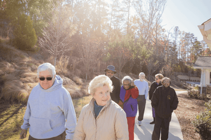 A group of seniors walks together along a sunny outdoor path surrounded by trees, enjoying an active lifestyle at an assisted living community in Mountainside, NJ.