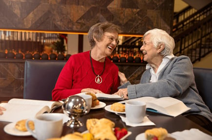 Two older adults share a warm, laughing moment over coffee and pastries in an elegant dining setting, reflecting the vibrant social atmosphere and resort-style senior living experience at Arbor Terrace Morris Plains, NJ.
