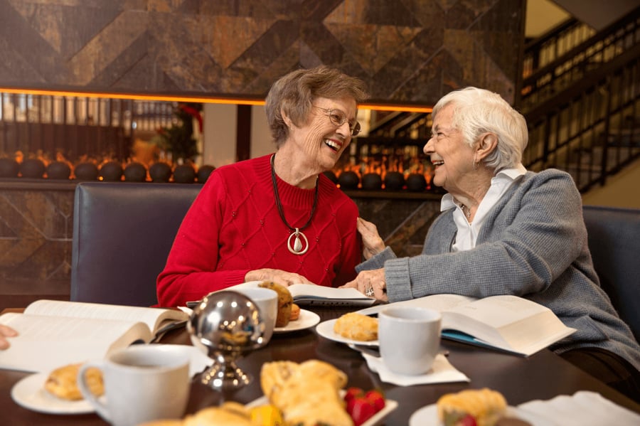 Two older adults share a warm, laughing moment over coffee and pastries in an elegant dining setting, reflecting the vibrant social atmosphere and resort-style senior living experience at Arbor Terrace Morris Plains, NJ.