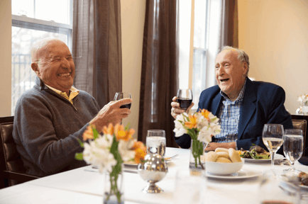 Two older adults laugh and clink glasses of wine at a beautifully set dining table with fresh flowers — warm dining options at senior living Lakeway Texas.