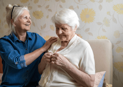 A warm moment of personalized assisted living in Woodbridge, VA, as a staff member gently helps an older adult resident with her clothing in a cheerfully wallpapered sitting area.