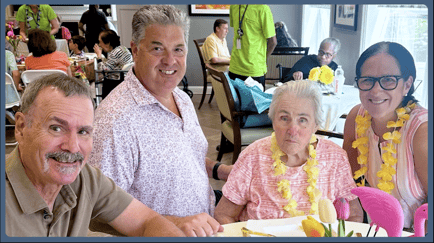 A senior woman wearing a yellow lei sits at a festively decorated table alongside smiling visitors during a themed dining event at Arbor Terrace Mountainside, reflecting the warm community spirit of this senior living community in Mountainside, NJ.