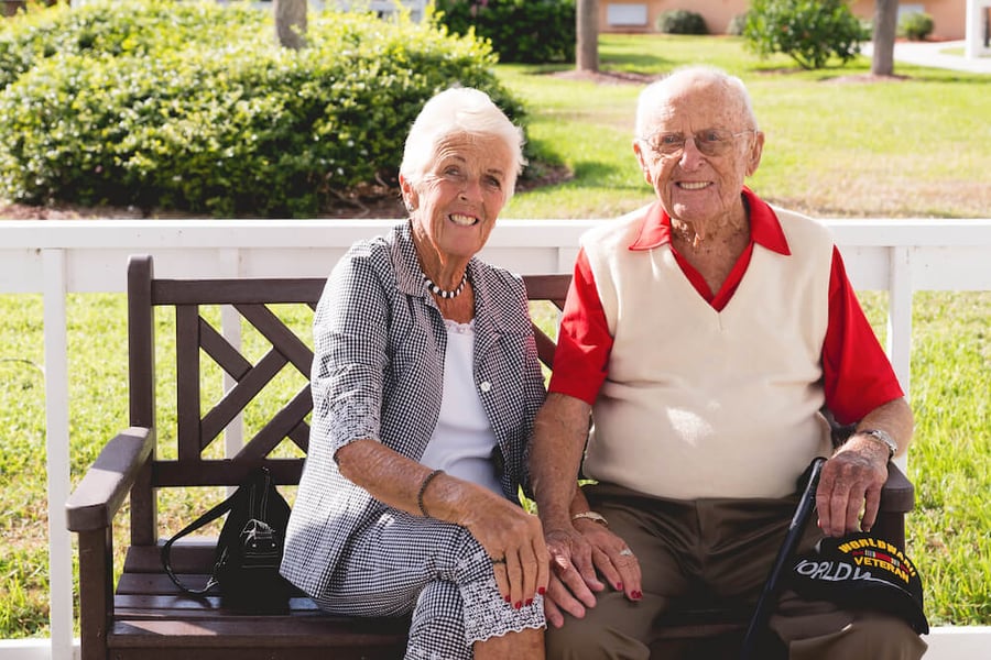 Two older adults sit side by side on a wooden park bench, smiling contentedly outdoors on a green lawn, enjoying the active senior lifestyle available in Fort Myers, FL.