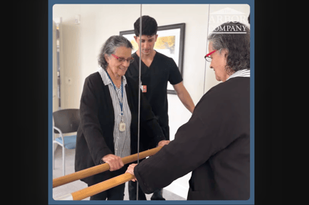 a senior lady holding on to a barre while a therapist assists her movements
