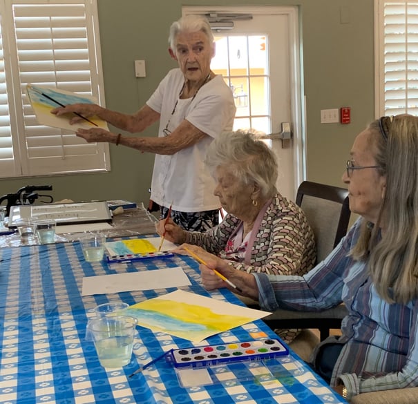 Two older residents concentrate on watercolor paintings during an art activity at Barrington Terrace of Fort Myers, FL, while another person holds up a finished canvas nearby in a bright, relaxed setting.