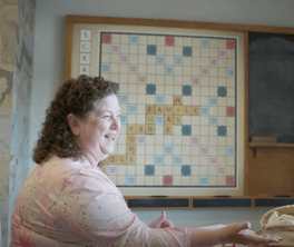 A woman with curly hair smiles mid-conversation in front of a wall-mounted word game board spelling out "family" at Arbor Terrace Prince William Commons, a lively senior living community in Woodbridge VA.