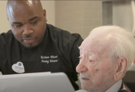 A dining director in a black chef's coat attentively reviews a menu on a tablet with an older male resident, highlighting the personalized assisted living Woodbridge VA experience and thoughtful senior care.