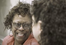 A woman with curly hair, glasses, and hoop earrings beams with delight during a face-to-face conversation at Arbor Terrace Prince William Commons, a joyful senior living community in Woodbridge VA.