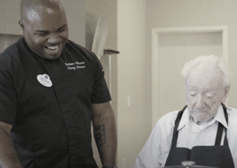 A dining team member laughs warmly alongside a resident wearing an apron during a cooking activity at Arbor Terrace Prince William Commons, a friendly senior living community in Woodbridge VA.