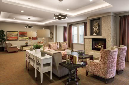 A beautifully furnished common living room featuring a stone fireplace, patterned armchairs, warm lighting, and vibrant artwork, offering a welcoming retreat at this boutique assisted living community in Woodbridge, VA.