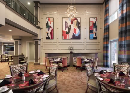 A sophisticated dining room with soaring ceilings, bold abstract artwork, a crystal chandelier, and elegantly set tables, showcasing the resort-style senior living experience in Woodbridge, VA.