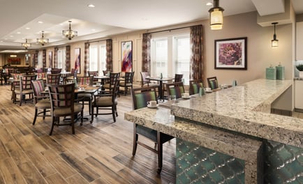 A spacious, elegantly appointed dining room with hardwood floors, granite countertops, pendant lighting, and colorful artwork, offering a warm restaurant-style setting at this senior living community in Woodbridge, VA.
