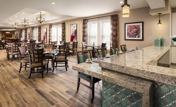 A spacious, elegantly appointed dining room with hardwood floors, granite countertops, pendant lighting, and colorful artwork, offering a warm restaurant-style setting at this senior living community in Woodbridge, VA.