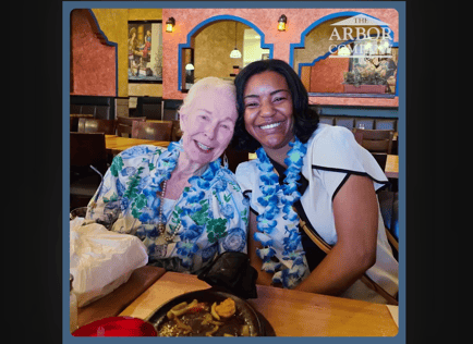 Two people beam with joy wearing matching blue floral leis at a festive restaurant outing, reflecting the fun, community-centered lifestyle and genuine bonds found at our senior living East Cobb and senior care Marietta GA community.
