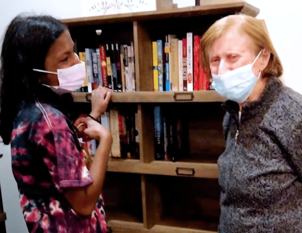 Two people wearing face masks share a warm interaction in front of a well-stocked bookshelf, reflecting the caring connections at assisted living in Suwanee, GA.