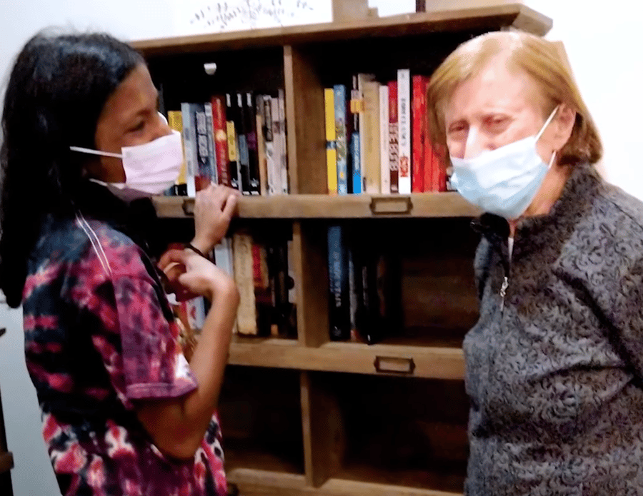 Two people wearing face masks share a warm interaction in front of a well-stocked bookshelf, reflecting the caring connections at assisted living in Suwanee, GA.
