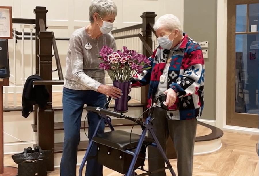 Two residents admire a vibrant bouquet of purple flowers in the warmly decorated entryway at Arbor Terrace Morris Plains, reflecting the caring, community-centered assisted living experience in Morris Plains, NJ.