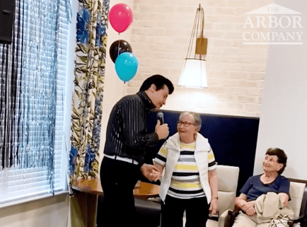 A performer in a sparkly jacket serenades a delighted resident with a microphone as others look on and smile at a festive balloon-filled event, showcasing the lively entertainment at our senior living East Cobb and assisted living Marietta GA community.